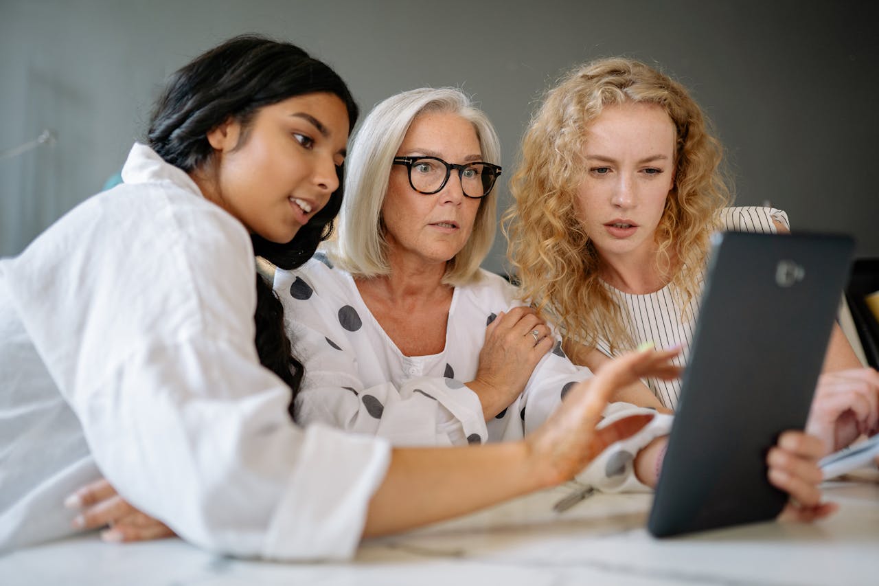 três mulheres olham a tela de um computador, sendo duas jovens e uma idosa