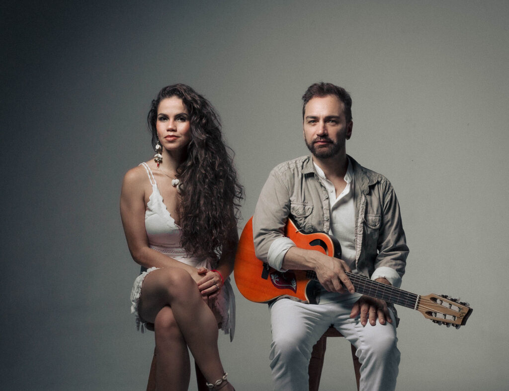 Casal, ela de vestido branco e cabelos compridos e ele de camisa cinza com violão vermelho