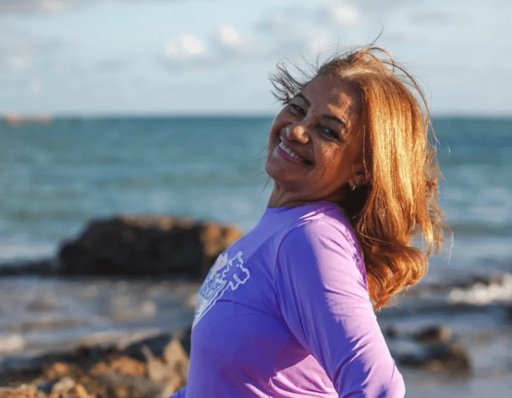 Mulher posa de perfil para a foto vestindo camiseta roxa a beira da praia