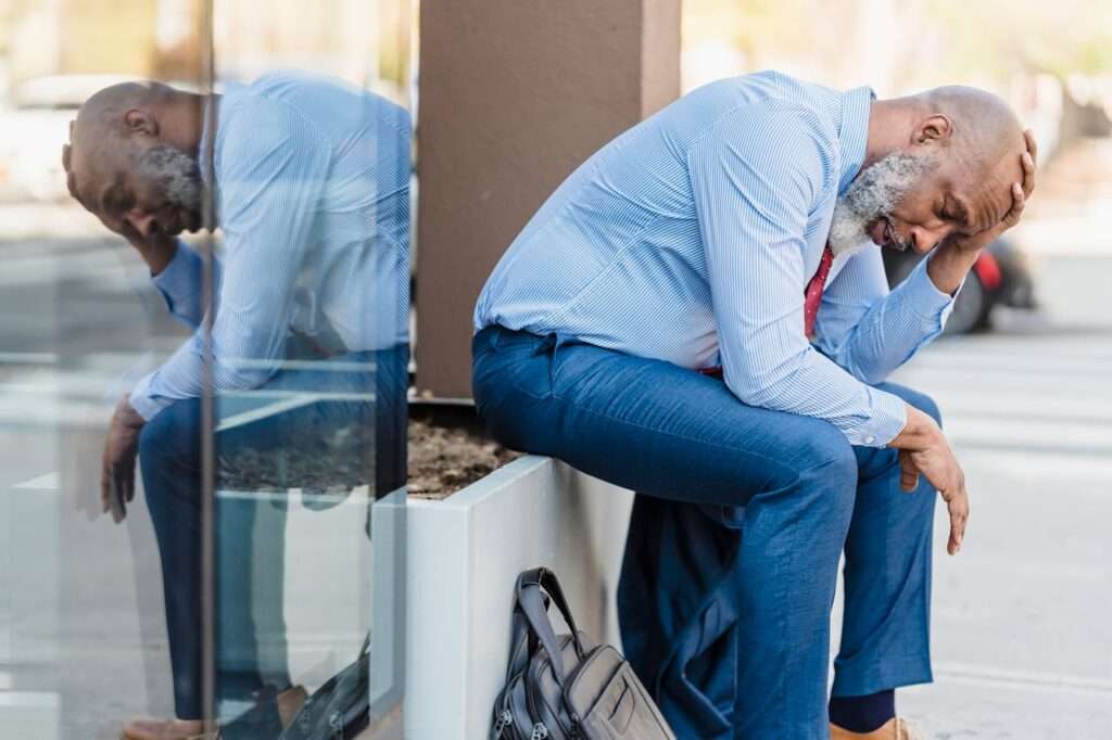 idoso negro, barba branca, camisa azul, está sentado e pensativo.