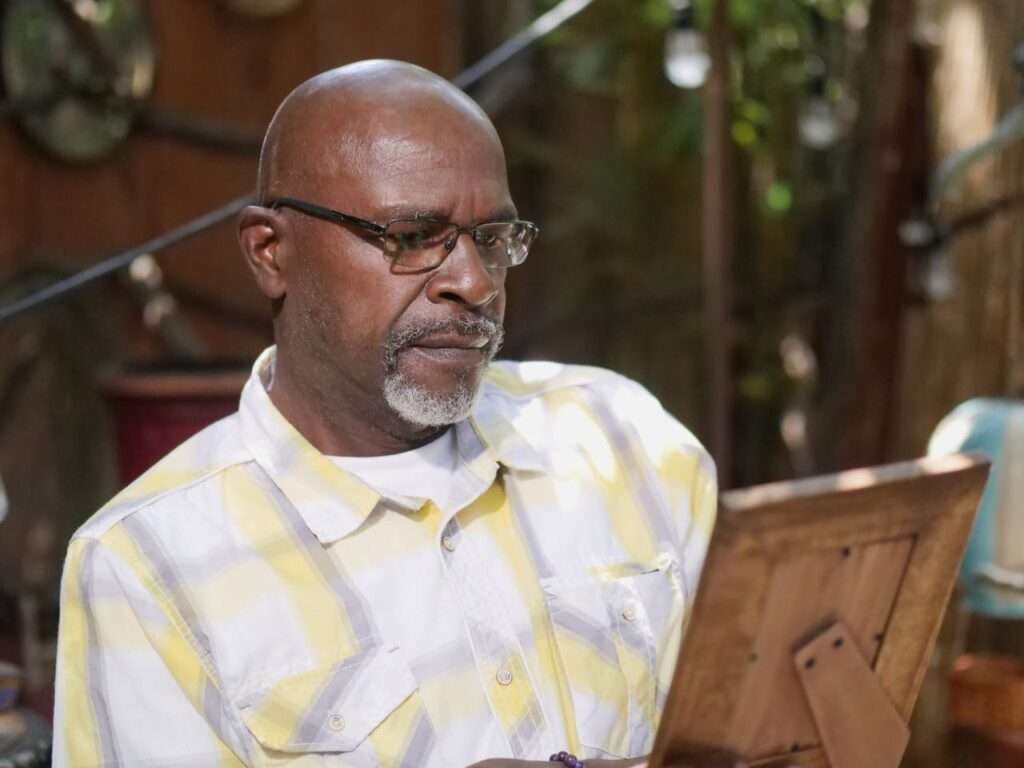 homem negro de barba branca e oculos e camisa branca e amarela olha um porta retrato