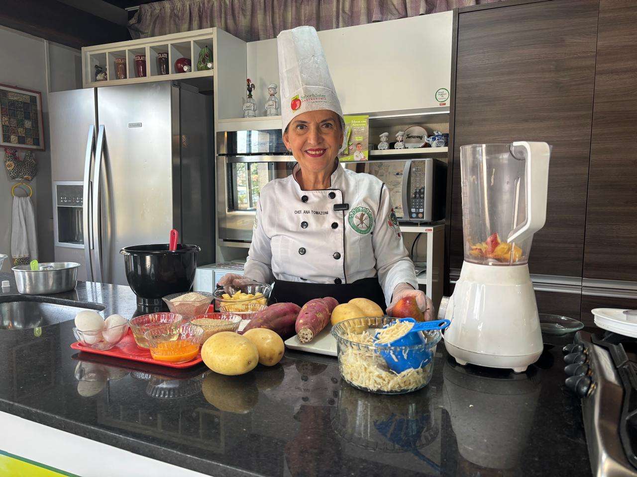 Mulher idosa vestida de chef em frente bancada de cozinha com ingredientes
