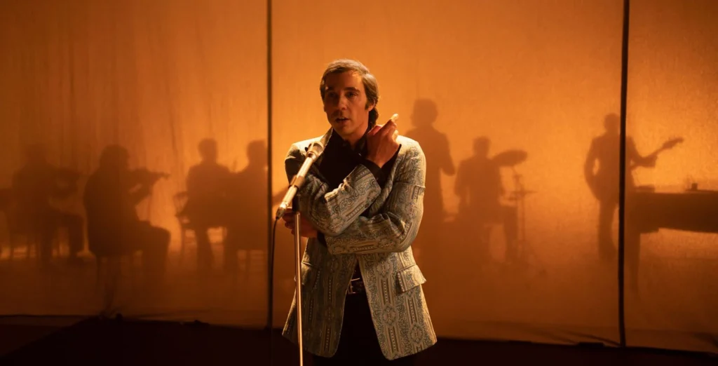 homem adulto canta no palco com luz amarronzada de fundo