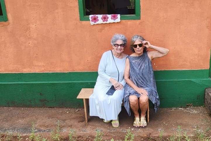 Duas idosas de óculos escuros sentadas em um banco, tendo ao fundo uma parede laranja e parte de janela verde